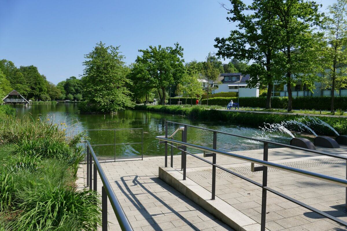Eine barrierefreie Rampe neben einem Teich mit fließendem Wasser, umgeben von Bäumen und Grünflächen an einem sonnigen Tag.