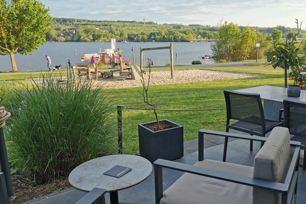 Von den Sitzgelegenheiten auf der Terrasse hat man an einem sonnigen Tag einen Blick auf einen See, einen Spielplatz und viel Grün.