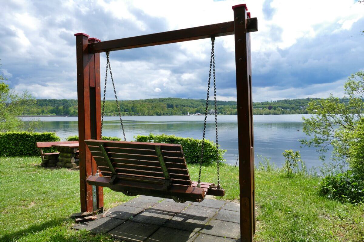 Eine Holzschaukel mit Blick auf den Möhnesee, im Hintergrund üppig grüne Bäume und ein bewölkter Himmel.