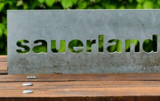 Metallschild mit dem ausgeschnittenen Schriftzug „Sauerland“, auf einer Holzunterlage platziert, im Hintergrund ist Grün zu sehen.
