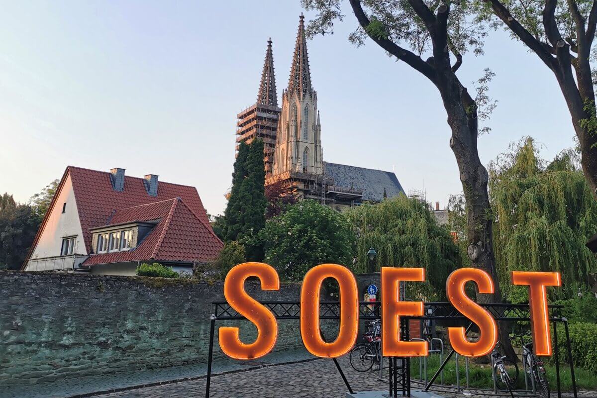 Großes, beleuchtetes SOEST-Schild vor historischen Kirchtürmen und Häusern, mit Bäumen und einer Steinmauer.