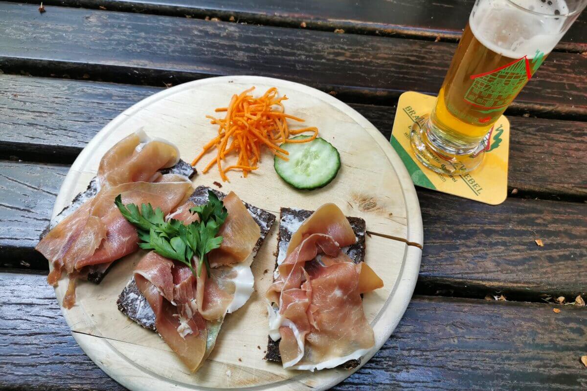 Pumpernickel mit Schinken, geraspelten Möhren, einer Gurkenscheibe und einem Glas Bier auf einem Holztisch.